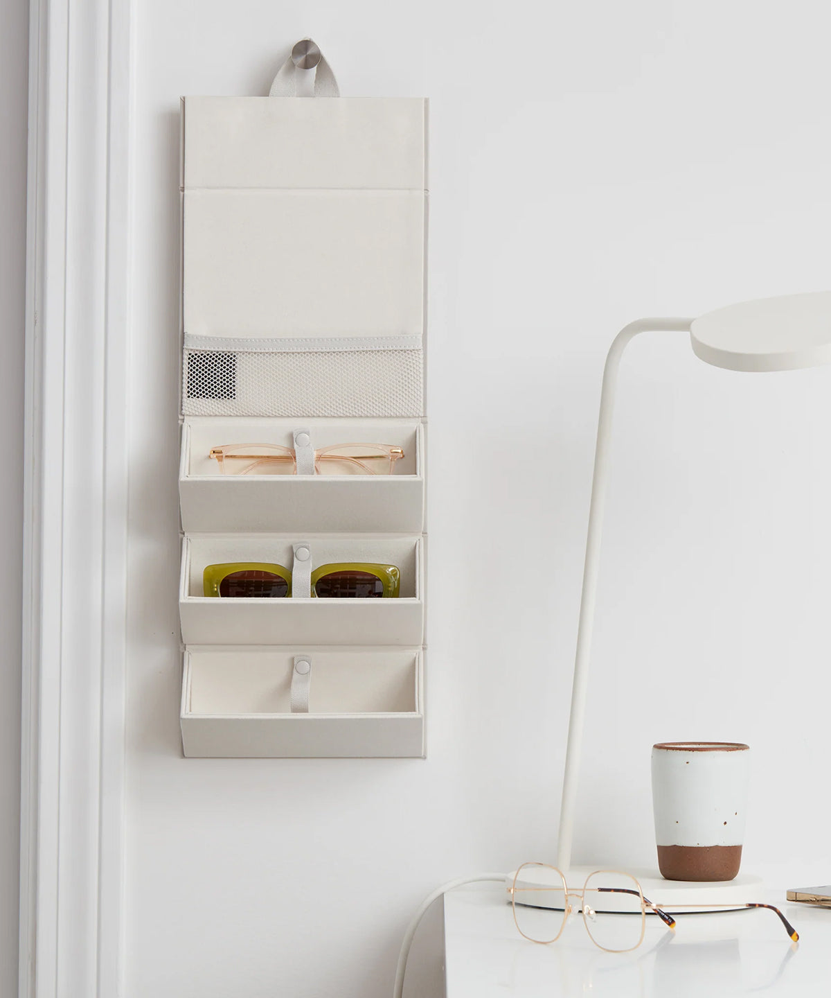 Dagne Dover Remi foldable glasses holder in white hung up and holding glasses by a desk.