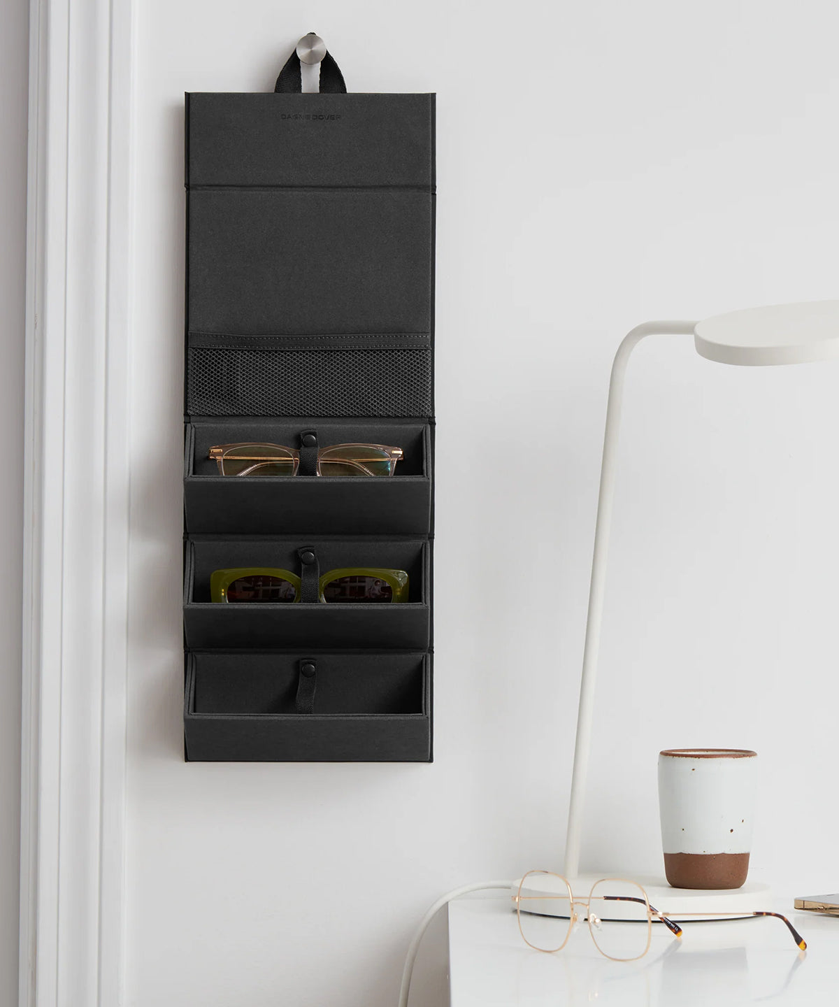 Dagne Dover Remi foldable glasses holder in black hung up and holding glasses by a desk.